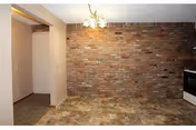 Empty room with a brick accent wall, tiled floor, and a hanging light fixture. There is a small open doorway on the left side leading to another area, and a white stove is partially visible on the right side.