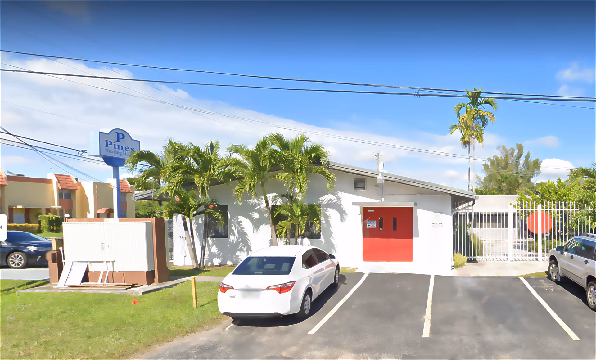 Exterior view of Pines Nursing Home building with a white car parked in front. The building is white with a red door and surrounded by palm trees. A sign with the facility name is visible on a pole near the street.