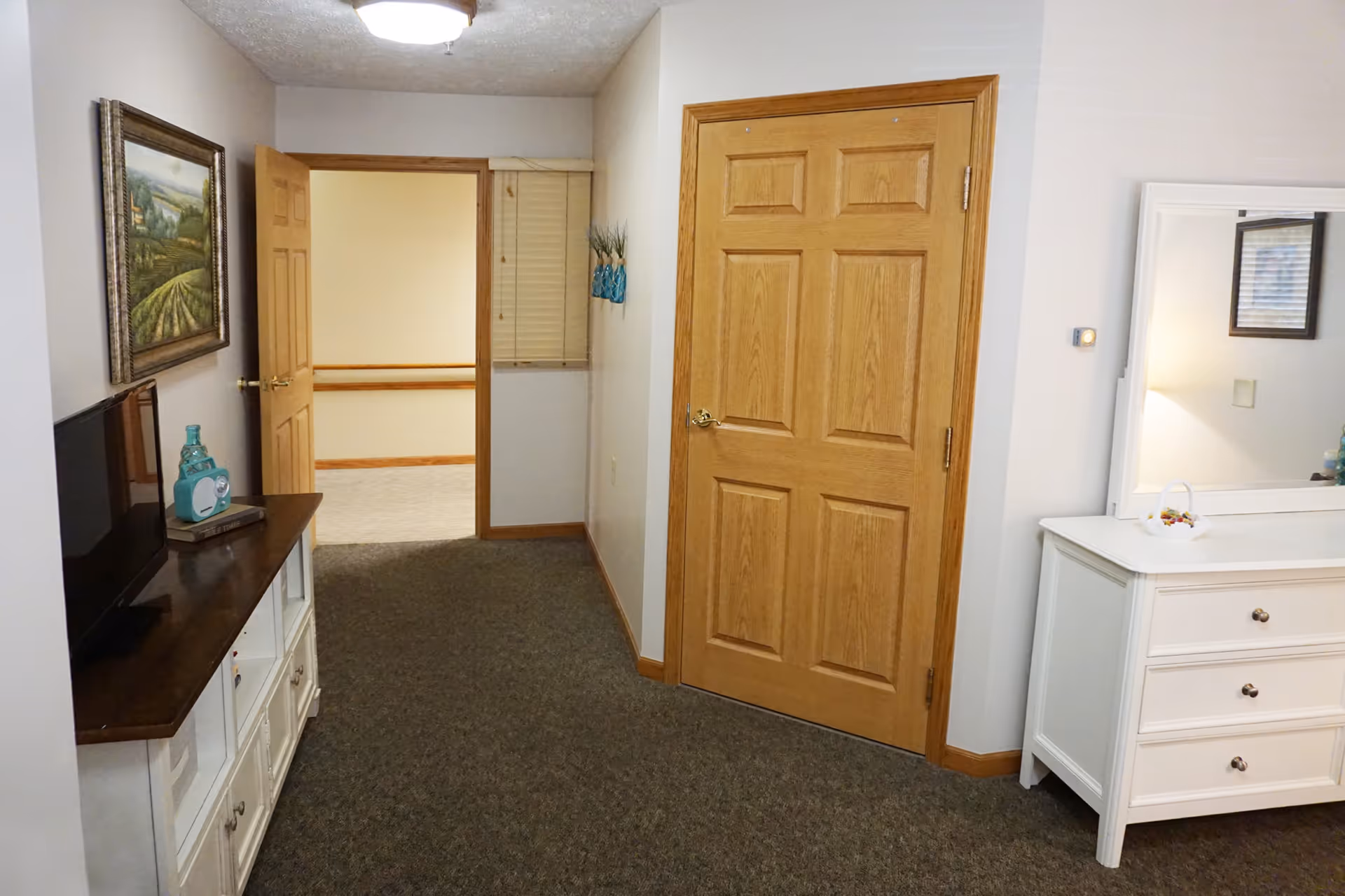 Hallway-style interior space with a closed wooden door, white dresser and mirror, TV stand, and an open doorway.