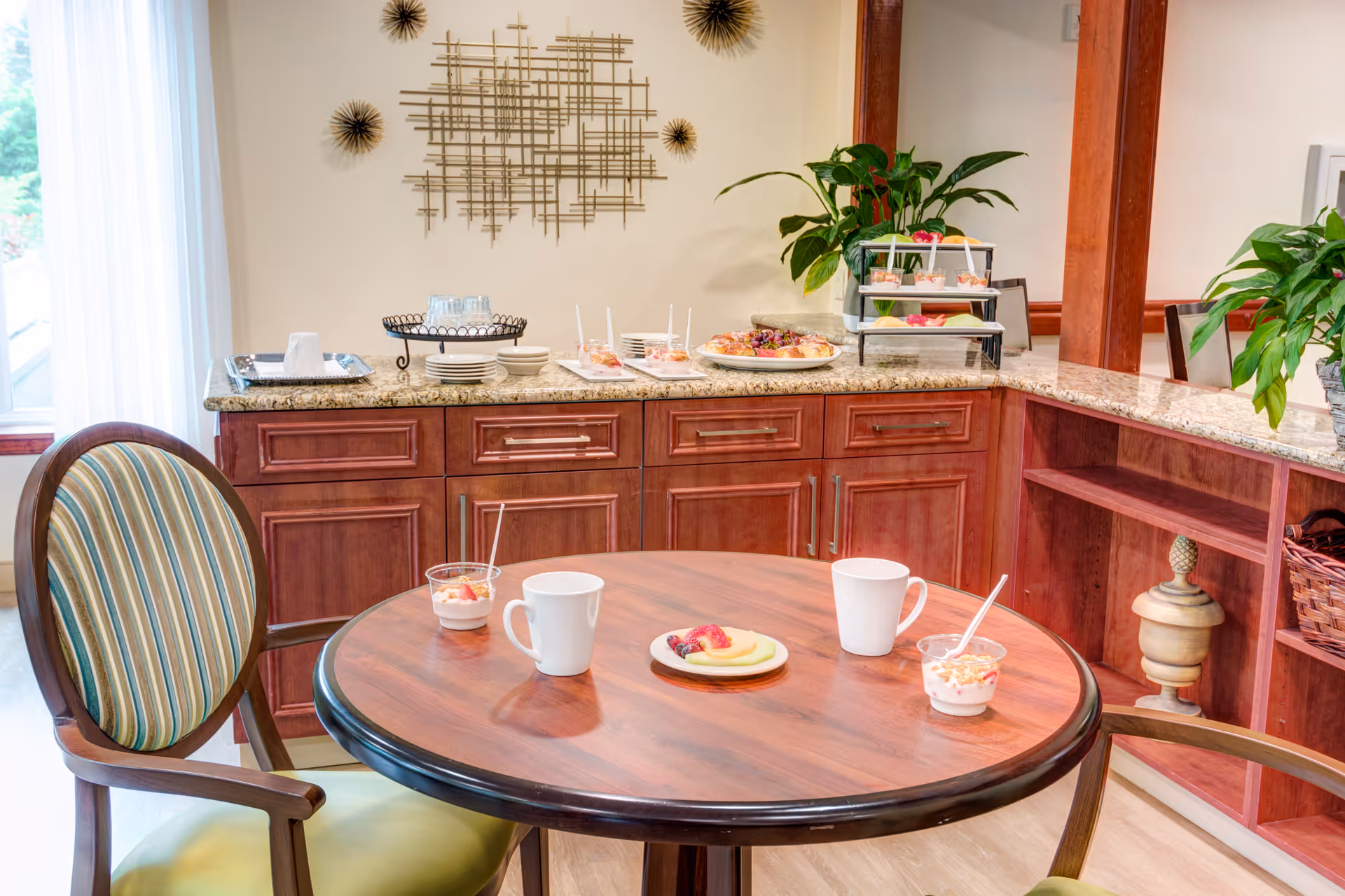 A cozy dining area with a round wooden table set with two white mugs, two cups of yogurt with spoons, and a small plate of sliced fruit. Behind the table is a granite countertop with wooden cabinets underneath, decorated with plates, glasses, trays of food, and green potted plants. A decorative metal wall art piece is mounted on the wall above the countertop.