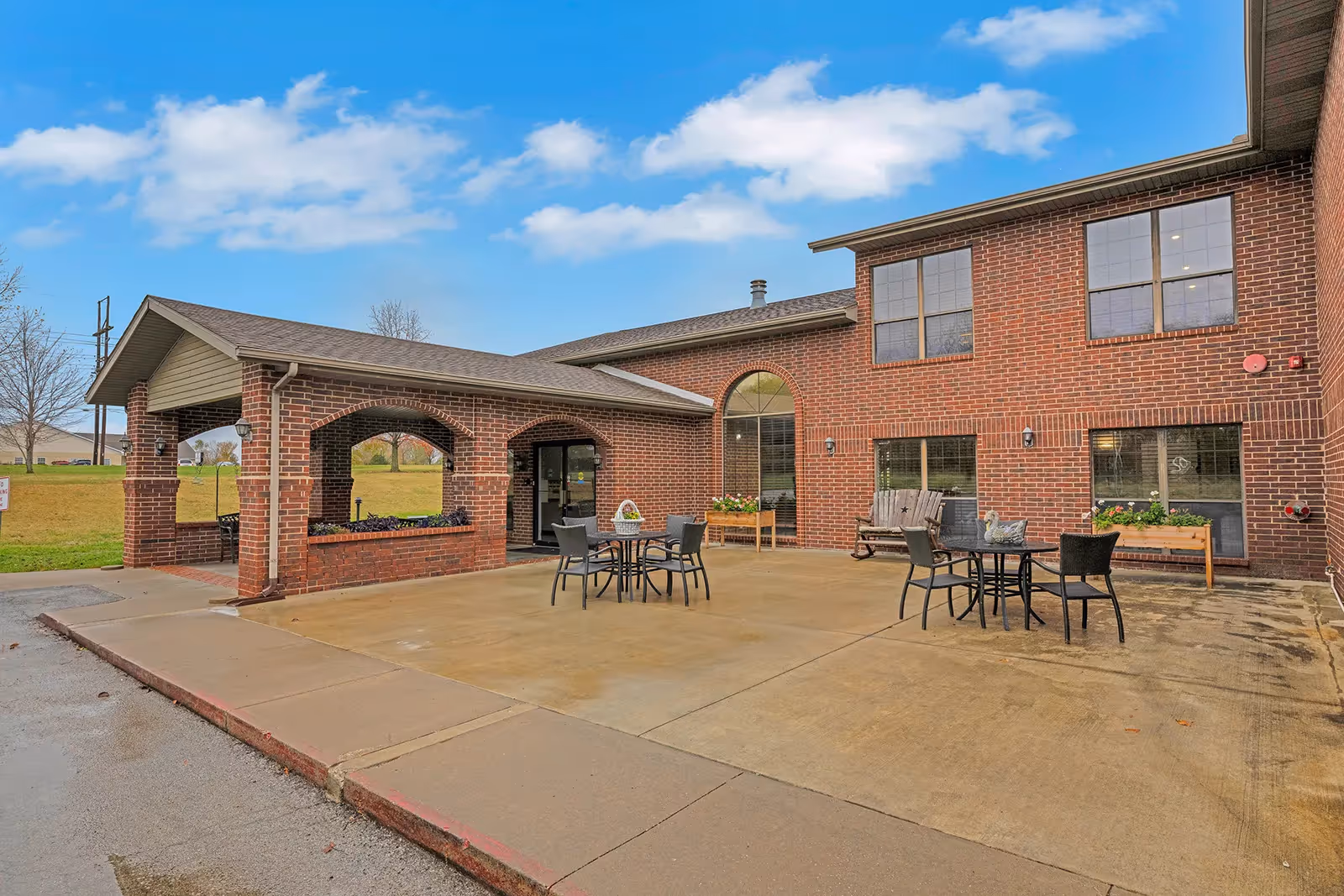 Outdoor patio area at The Bungalows at Nevada featuring a brick building with large windows, several round tables with chairs, and flower planters. The sky is blue with some clouds.