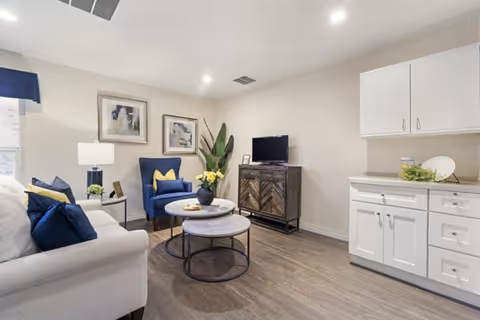 A cozy living room with a white sofa adorned with blue and yellow pillows, a blue armchair with a yellow pillow, two round nested coffee tables with a vase of yellow flowers, a wooden TV stand with a flat-screen TV, two framed abstract paintings on the wall, a tall green plant in the corner, and white cabinetry on the right side. The room has wooden flooring and recessed ceiling lights.
