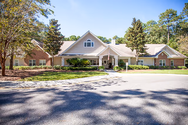 Front exterior view of Harbor Cove Memory Care facility, a single-story building with a combination of brick and siding, a central entrance with a covered porch, surrounded by trees and landscaping under a clear blue sky.