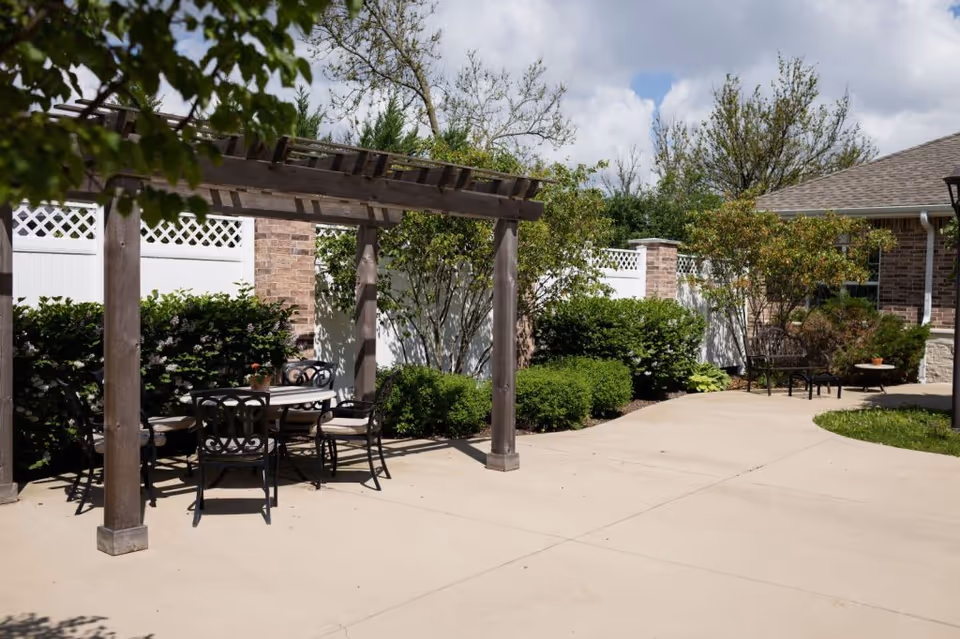 Outdoor patio area with a wooden pergola covering a round table and four chairs. The patio is surrounded by green bushes, trees, and a white fence with brick pillars. A building with brick and stone exterior is visible in the background under a partly cloudy sky.