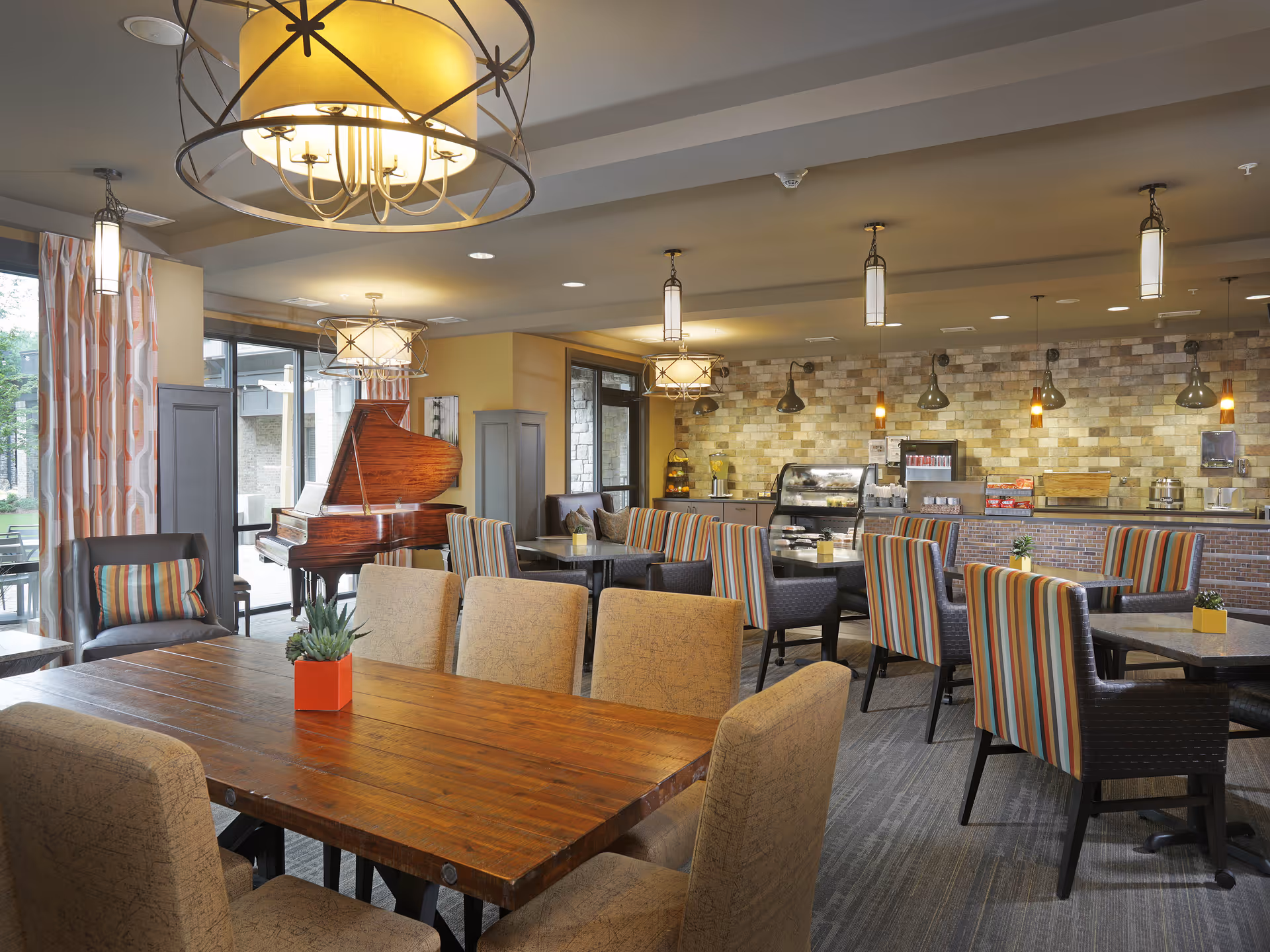 A spacious dining area in a senior living facility with a large wooden table surrounded by beige upholstered chairs in the foreground. In the background, there are multiple tables with striped chairs and a buffet counter with various food and drink items. The room features modern hanging light fixtures, a brick accent wall, large windows with patterned curtains, and a grand piano near the windows.