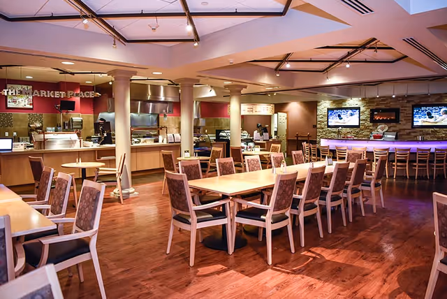 A spacious dining area with wooden floors and multiple tables and chairs arranged neatly. In the background, there is a food service counter labeled 'The Market Place' with kitchen equipment and a staff member. On the right side, there is a bar area with high chairs and two wall-mounted televisions above a stone accent wall.