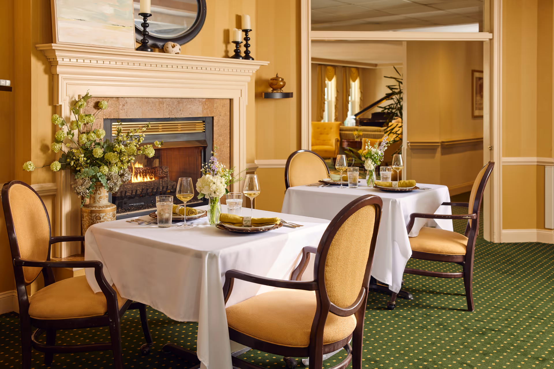 A cozy dining area with two tables covered in white tablecloths, each set with plates, glasses, and napkins. Four upholstered chairs with wooden frames surround the tables. A lit fireplace with a decorative mantel is in the background, adorned with a vase of flowers, candles, and a round mirror above it. The room has warm yellow walls and green carpet with a dotted pattern. A doorway leads to another room with a piano and yellow armchairs.