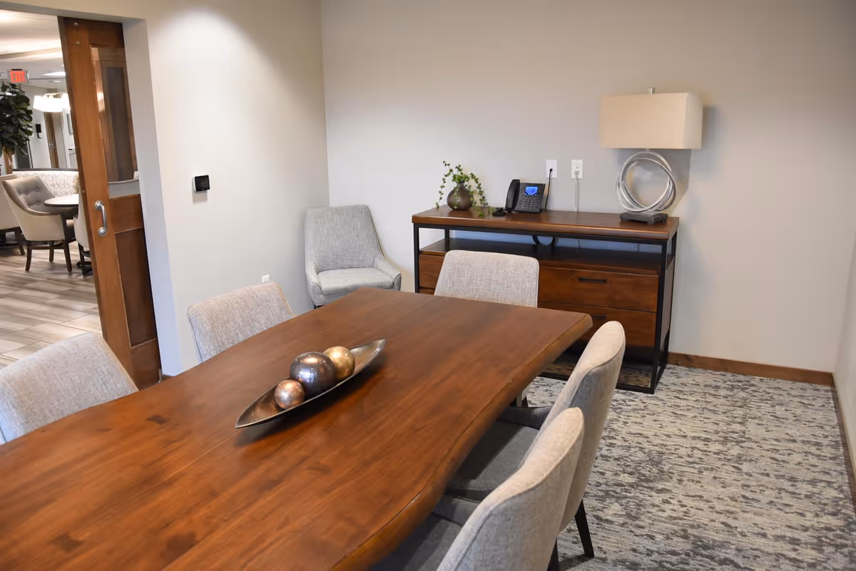 Small dining/meeting room with a wooden table surrounded by upholstered chairs and a sideboard holding a lamp and phone.