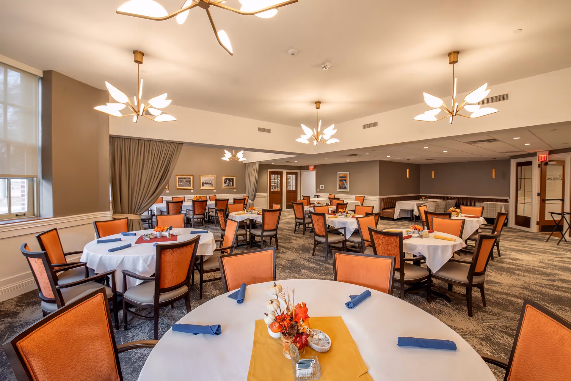 A spacious dining room with multiple round tables covered with white tablecloths and orange chairs. Each table is set with blue napkins and small autumn-themed centerpieces. The room features modern light fixtures, large windows with curtains, and framed pictures on the walls.