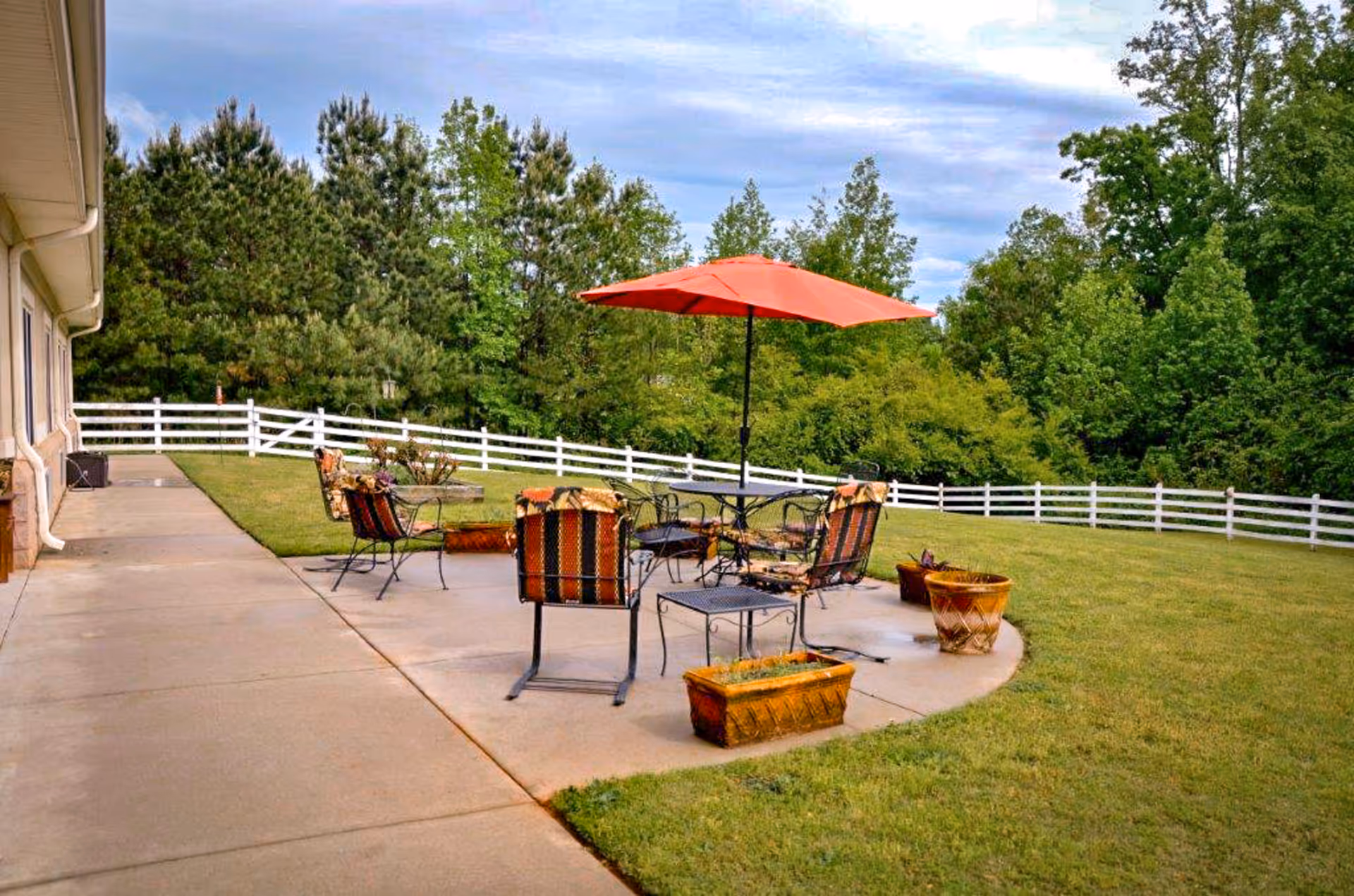Outdoor patio area with metal chairs and tables, some with patterned cushions, and a large red umbrella. The patio is surrounded by a grassy lawn, potted plants, a white wooden fence, and trees in the background under a partly cloudy sky.