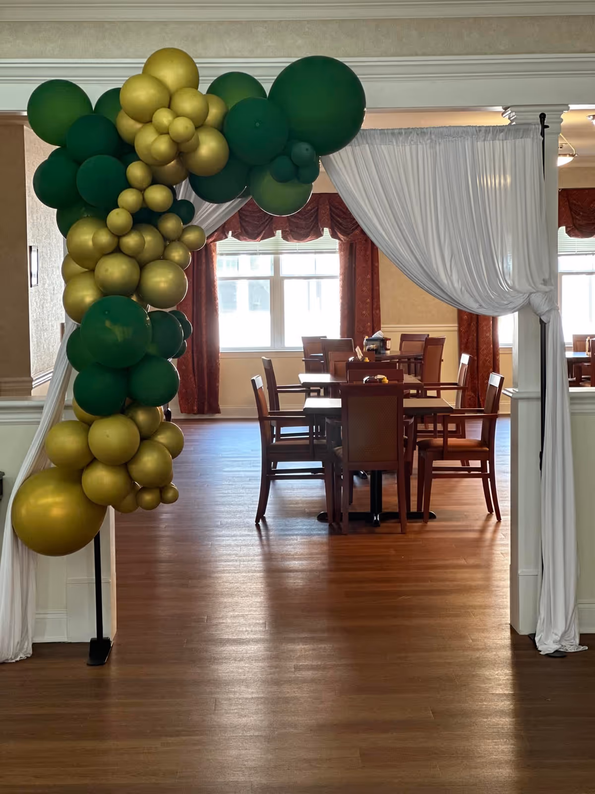 An interior dining area decorated with a green and gold balloon arch on the left side. The room has wooden floors, several tables with chairs, and large windows with red curtains letting in natural light. White drapes hang from the ceiling framing the entrance to the dining space.