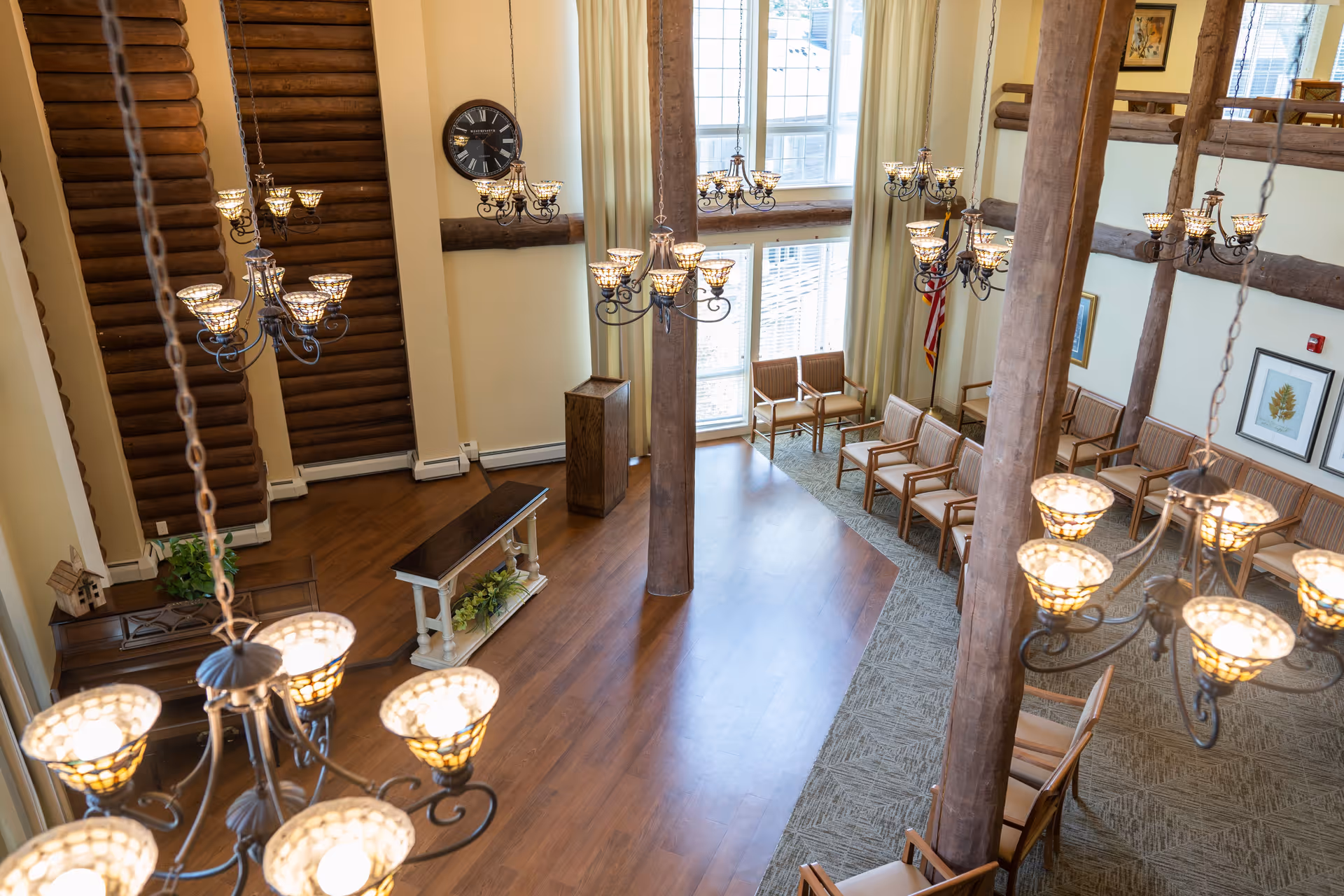 Interior view of a spacious common area with high ceilings, wooden beams, multiple chandeliers, a clock on the wall, and rows of wooden chairs arranged along the carpeted and hardwood floors near large windows with curtains.