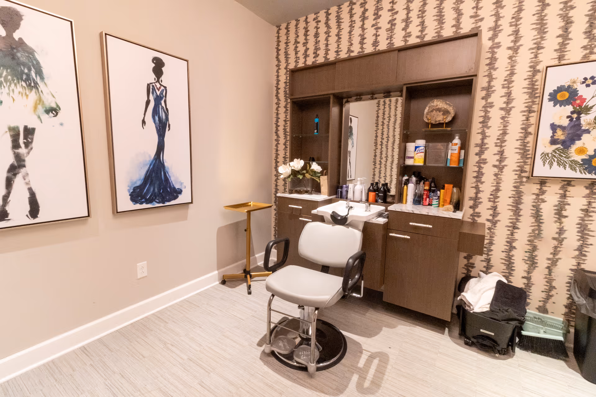 A salon styling station with a barber chair, wash sink, mirror and cabinets, flanked by framed artwork and patterned wallpaper.