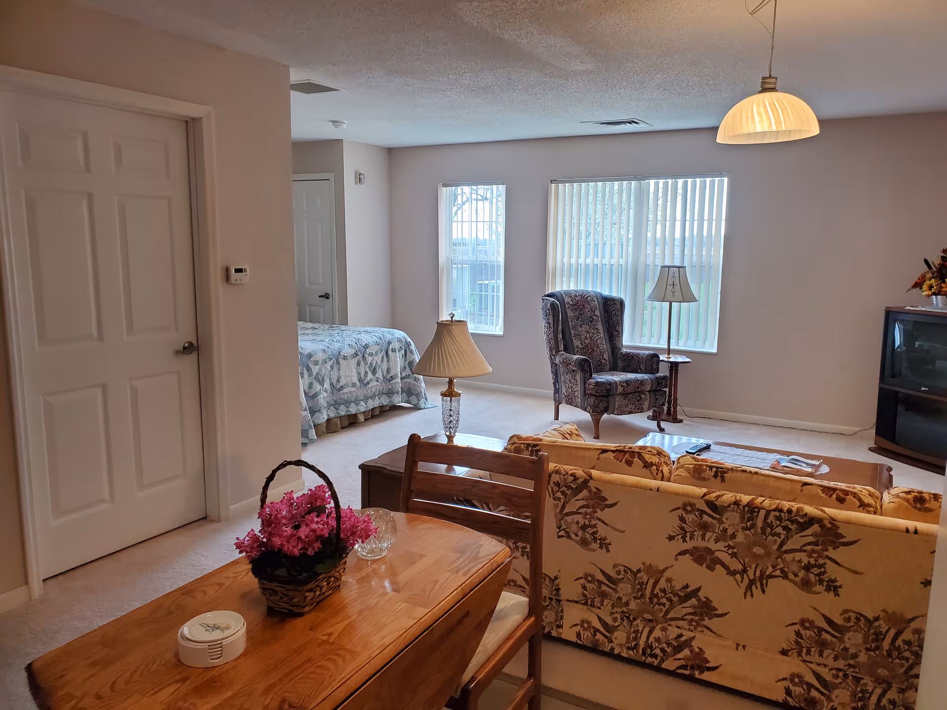 A cozy living area in a senior living facility with a floral patterned sofa, wooden coffee table, and a floral armchair near two windows with vertical blinds. A bed with a quilted bedspread is visible in an adjacent bedroom. The room is softly lit with a hanging ceiling lamp and a table lamp on a side table. A TV is placed in the corner on a stand.