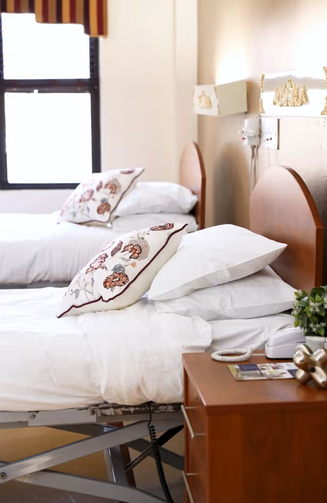 Two single beds with white linens and floral pillows in a bright senior care bedroom with a bedside table and phone.