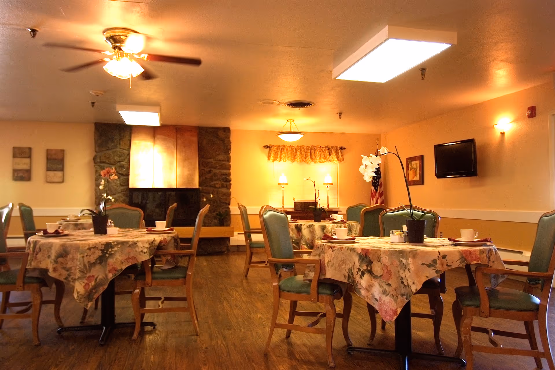 A cozy dining room in a nursing home with round tables covered in floral tablecloths, each set with cups and plates. Green cushioned wooden chairs surround the tables. The room features a stone fireplace with a copper hood, a ceiling fan, wall-mounted TV, and warm lighting. There are decorative plants on the tables and framed artwork on the walls.