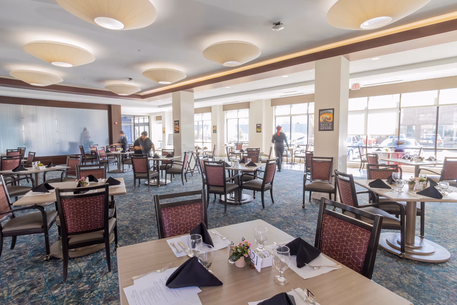 A bright and spacious dining room with multiple tables set with black napkins, glasses, silverware, and small flower arrangements. The room has large windows allowing natural light to fill the space, patterned carpet flooring, and modern ceiling lights. Two staff members are seen attending to the tables.