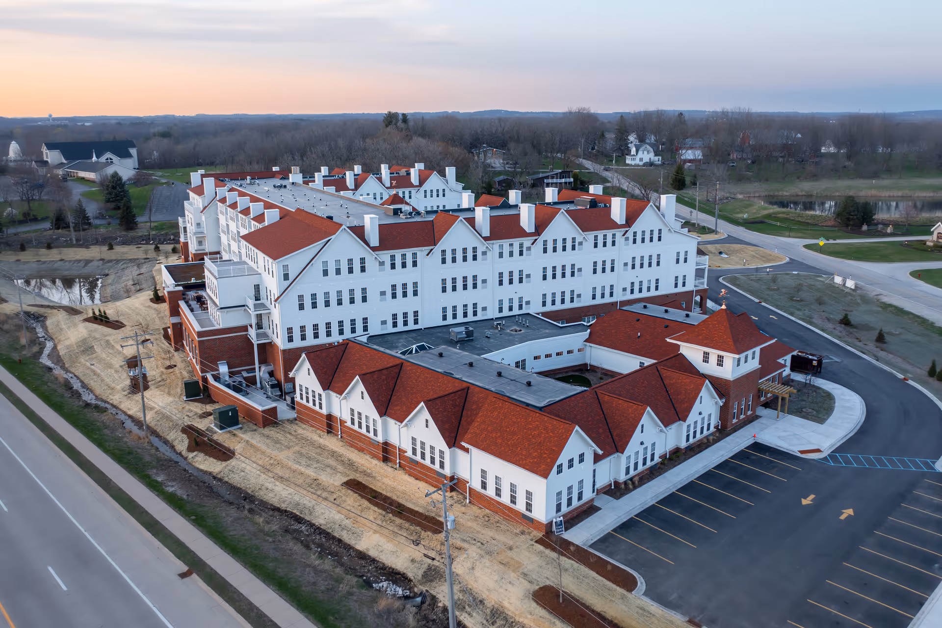 Aerial view of a large white multi-story senior living building with red roofs, parking lot, and surrounding roads.