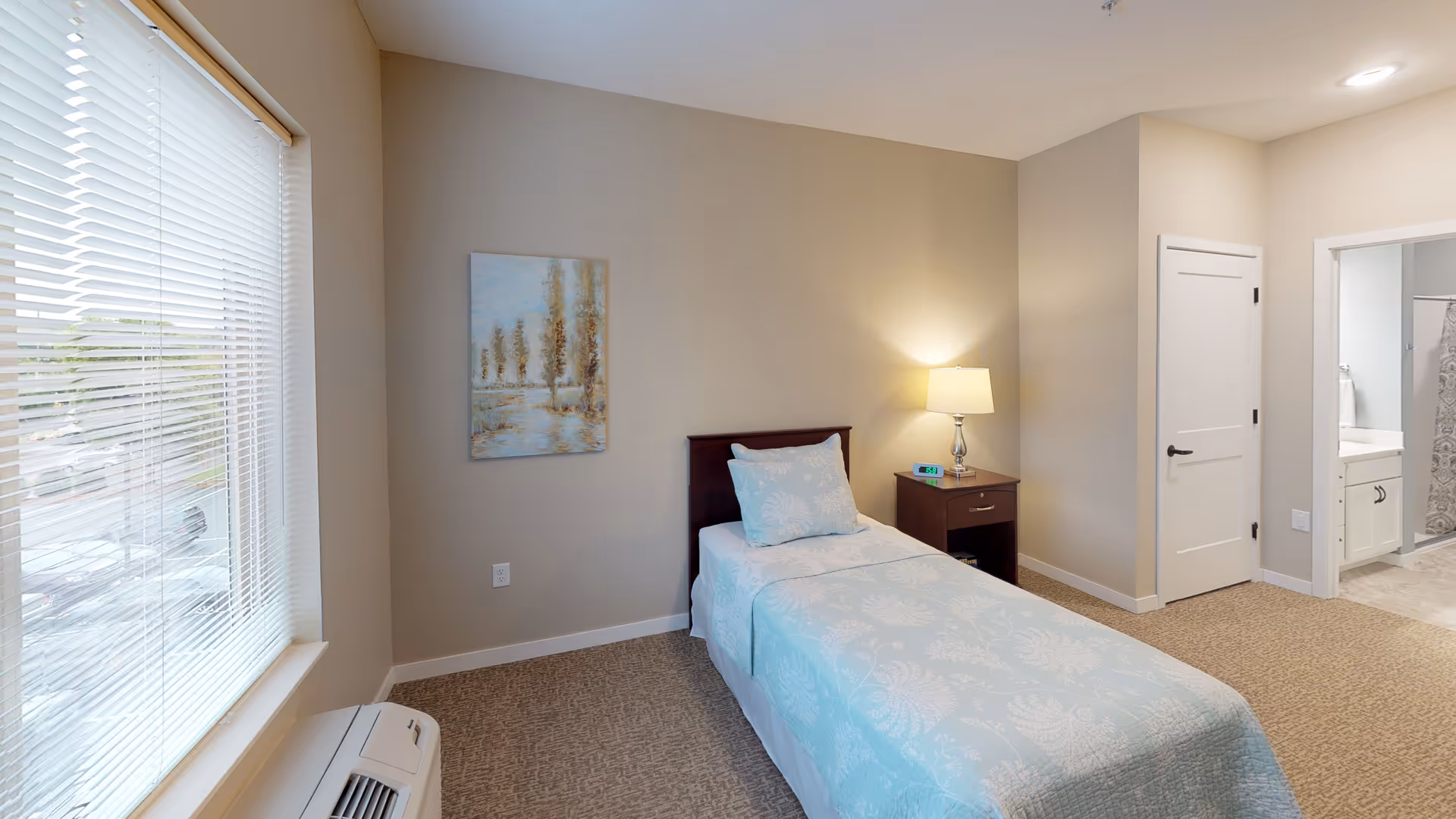 A single bedroom with a bed covered in light blue patterned bedding, a wooden nightstand with a lamp and alarm clock, beige walls, carpeted floor, a window with blinds, and a door leading to a bathroom.