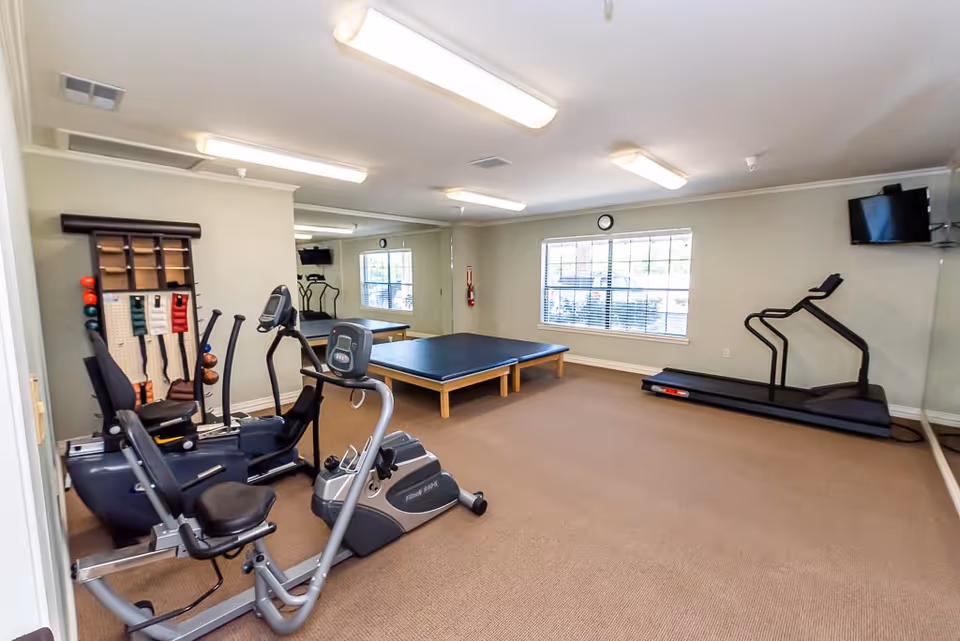 A fitness room in a senior living facility featuring exercise equipment including a recumbent bike, an elliptical machine, a treadmill, and a table for physical therapy or stretching. The room has beige carpet, light-colored walls, a large window with blinds, a wall-mounted TV, and fluorescent ceiling lights.
