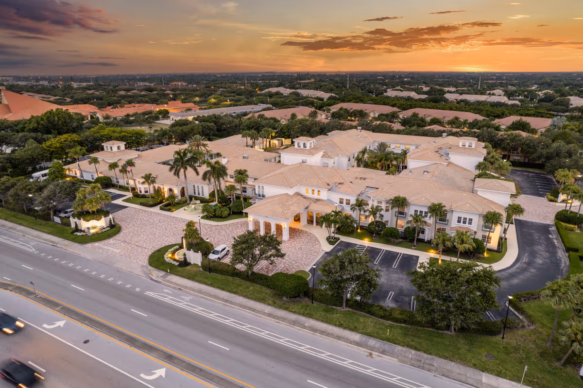 Aerial view of Tequesta Terrace senior living facility at sunset, showing a large, light-colored building with a tiled roof, surrounded by palm trees and landscaped grounds, with a parking area and a road in the foreground.