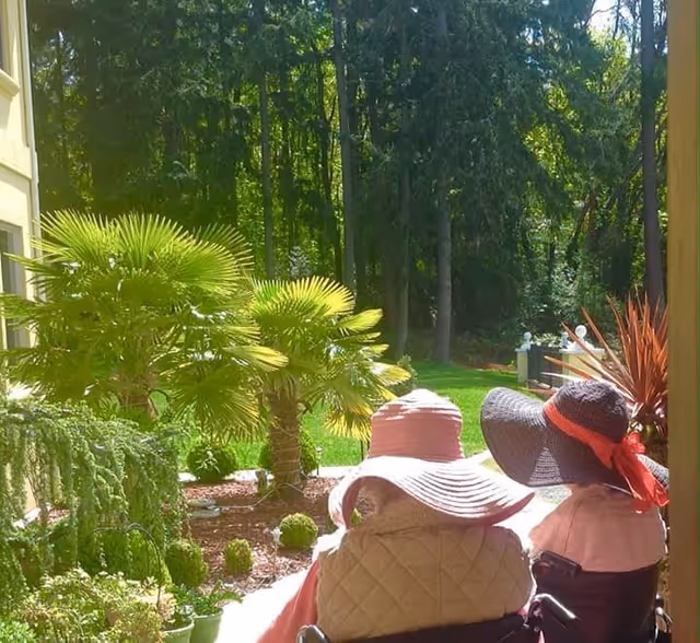 Two elderly individuals wearing large sun hats sitting outdoors in wheelchairs, facing a lush garden with palm trees and other greenery, enjoying a sunny day at Villa Vecchio at Dash Point Adult Family Home.