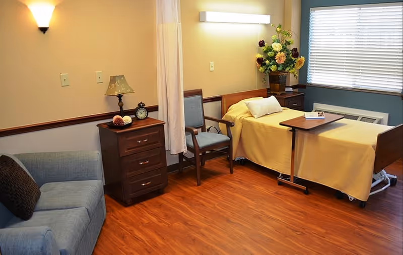A cozy senior living room with a hospital-style bed covered in a yellow blanket, a wooden bedside table with a lamp and clock, a cushioned armchair, a blue sofa with a dark pillow, a wooden floor, a window with blinds, and a vase of flowers on a small table near the bed.