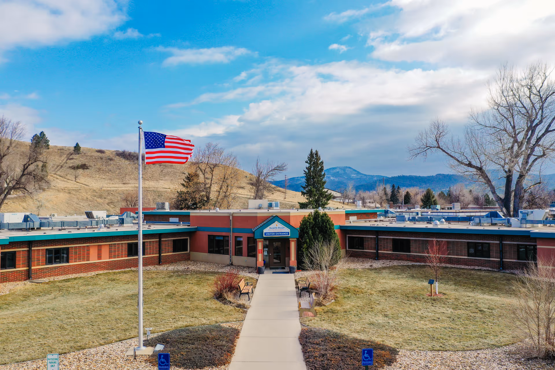 Front exterior of a single-story healthcare building with an American flag, central walkway, and mountains in the background.