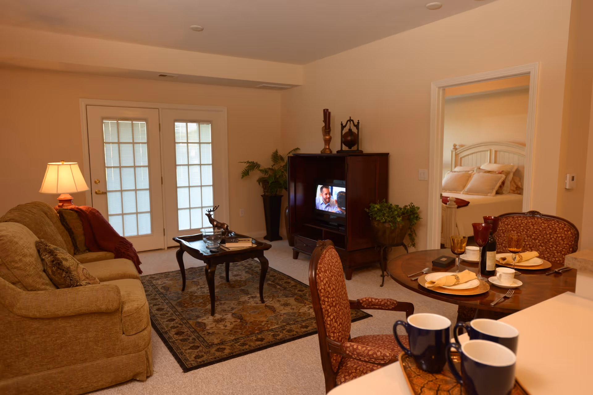 A cozy living room with a beige sofa, a wooden coffee table on a patterned rug, and a TV cabinet with a television showing a man. There is a round dining table set with plates, napkins, glasses, and a bottle of wine. A doorway leads to a bedroom with a neatly made bed. French doors with frosted glass panels are visible in the background.