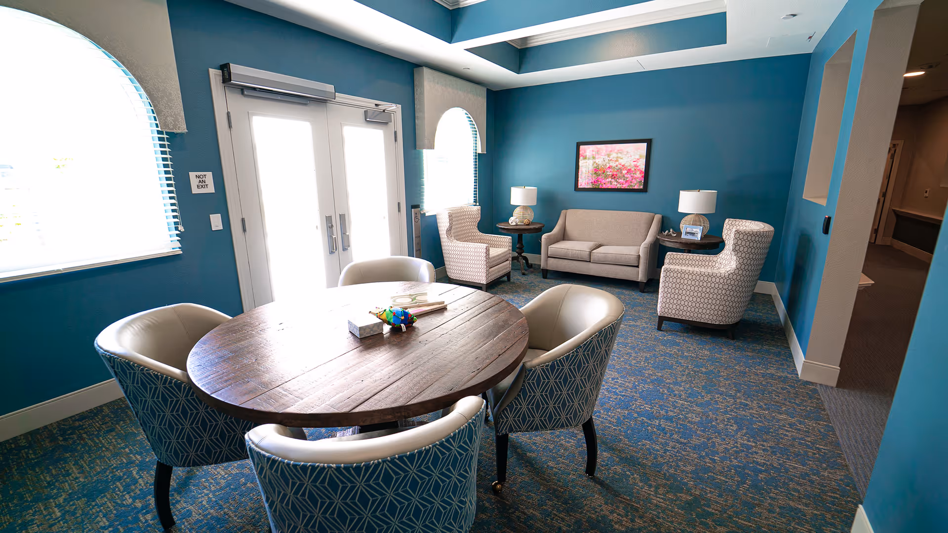 A cozy senior living common area with teal walls and patterned carpet. The room features a round wooden table surrounded by four upholstered chairs with geometric patterns. In the background, there is a beige sofa flanked by two matching armchairs and side tables with lamps. A framed floral picture hangs on the wall above the sofa. Two arched windows and a set of double doors allow natural light into the room.