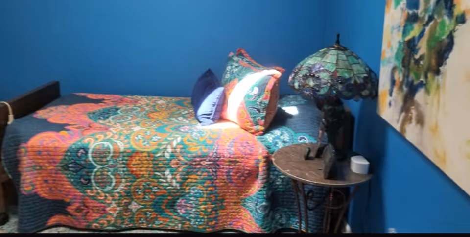 A bedroom with a bed covered in a colorful patterned quilt and several pillows. Next to the bed is a small round table with a decorative stained glass lamp and some small items. The walls are painted blue and there is a colorful abstract painting hanging on the wall.
