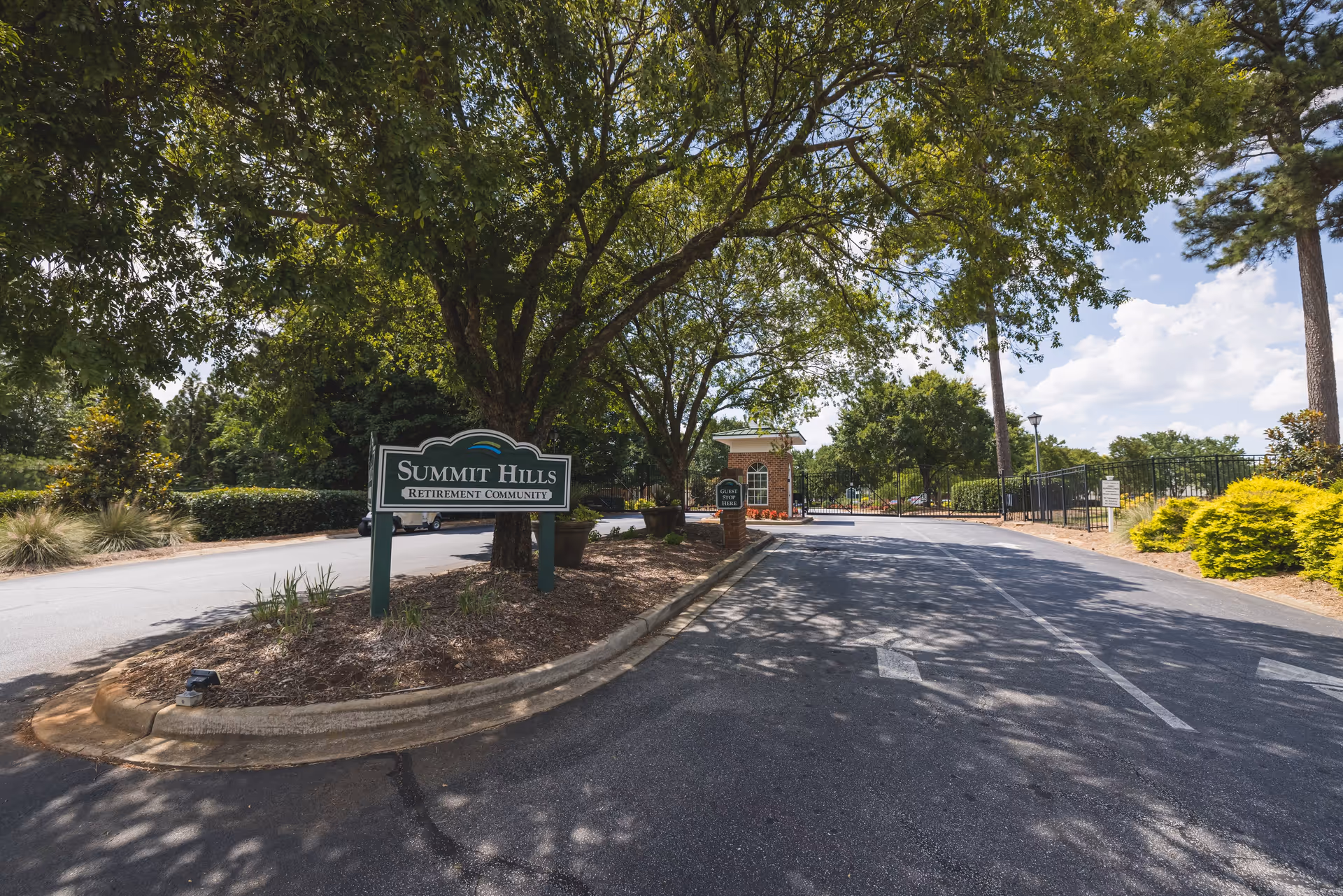 Entrance road to Summit Hills Retirement Community with a green sign on the left side surrounded by trees and landscaping, a gated security booth ahead, and a clear blue sky with some clouds.