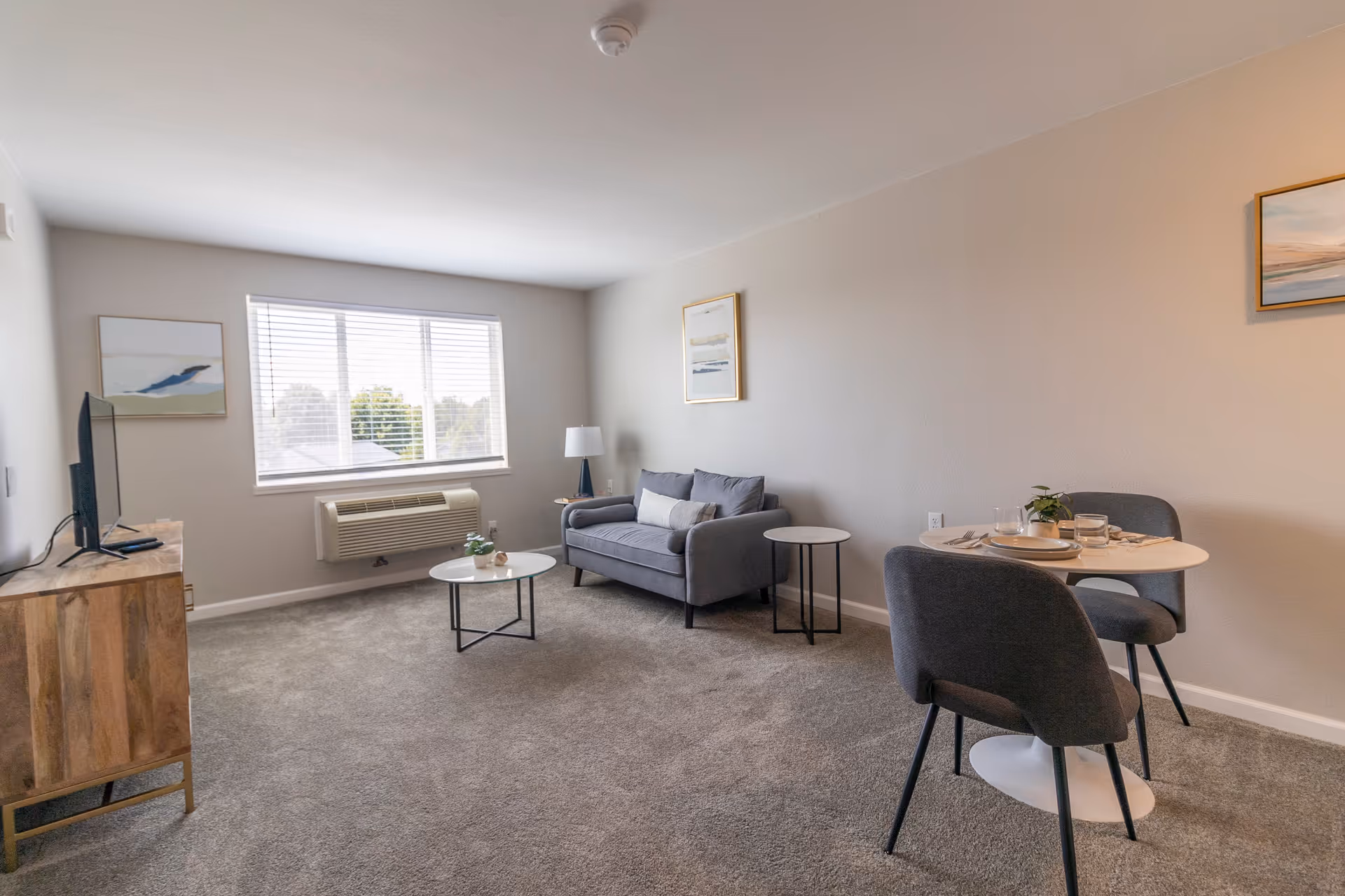 A bright living room with a large window letting in natural light. The room features a gray loveseat with a pillow, a small round coffee table, a side table with a lamp, a wooden TV stand with a flat-screen TV, and a small round dining table set with two gray chairs and tableware. The walls are light-colored with framed artwork.