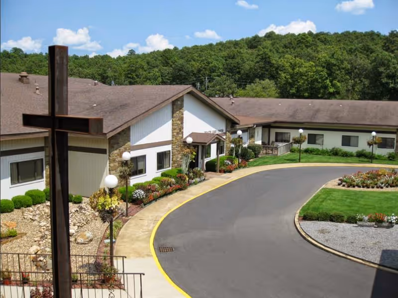 Entrance and curved driveway of a single-story senior living facility with landscaped gardens and a large wooden cross in the foreground.