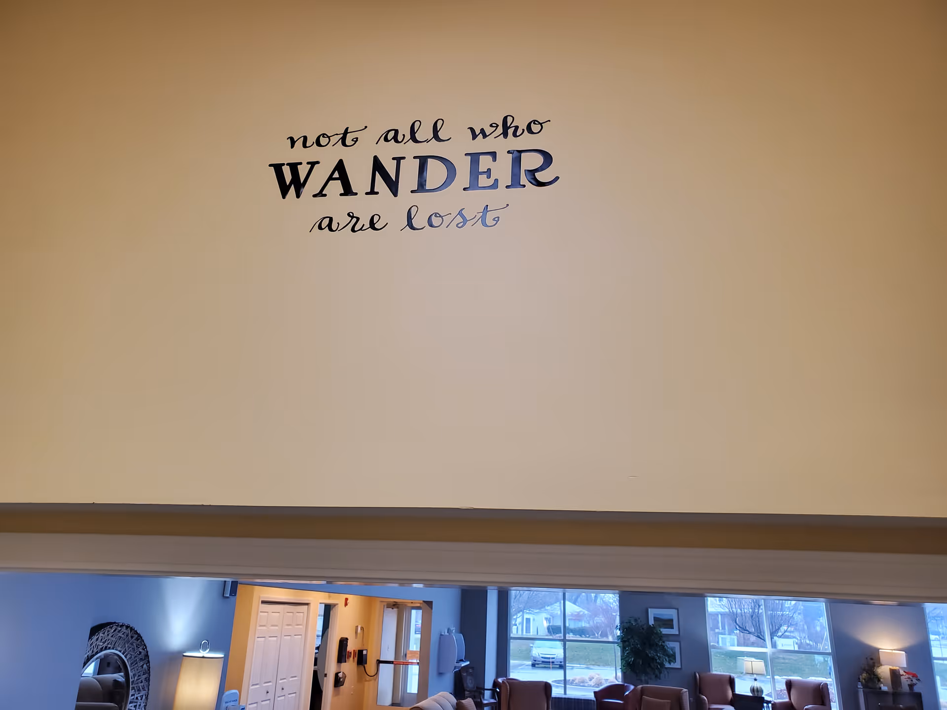 Interior view of a memory care community with a beige wall displaying the phrase 'not all who WANDER are lost' in decorative lettering above a doorway. Below the wall is a common area with chairs, a lamp, a mirror, and large windows showing an outdoor scene.