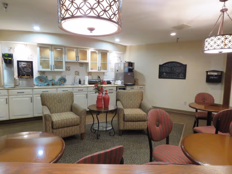 A cozy common area in an independent living facility featuring two patterned armchairs around a small round table with red vases and flowers. The background shows a kitchenette with white cabinets, a coffee machine, and a water dispenser. There are also round tables with red striped chairs and decorative hanging light fixtures.