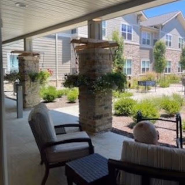 Covered outdoor patio area with cushioned chairs and a small table, overlooking a landscaped garden with shrubs and a multi-story senior living building in the background.