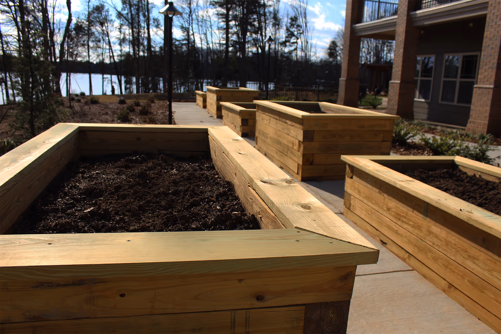 Outdoor garden area with multiple wooden raised garden beds filled with soil, situated on a paved walkway near a building and surrounded by trees with a lake visible in the background under a partly cloudy sky.