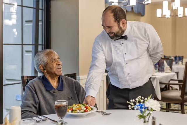 An elderly man sitting at a dining table with a glass of iced tea and a plate of salad in front of him, looking up and smiling at a waiter dressed in a white shirt and black bow tie who is serving the salad to him in a well-lit dining room.