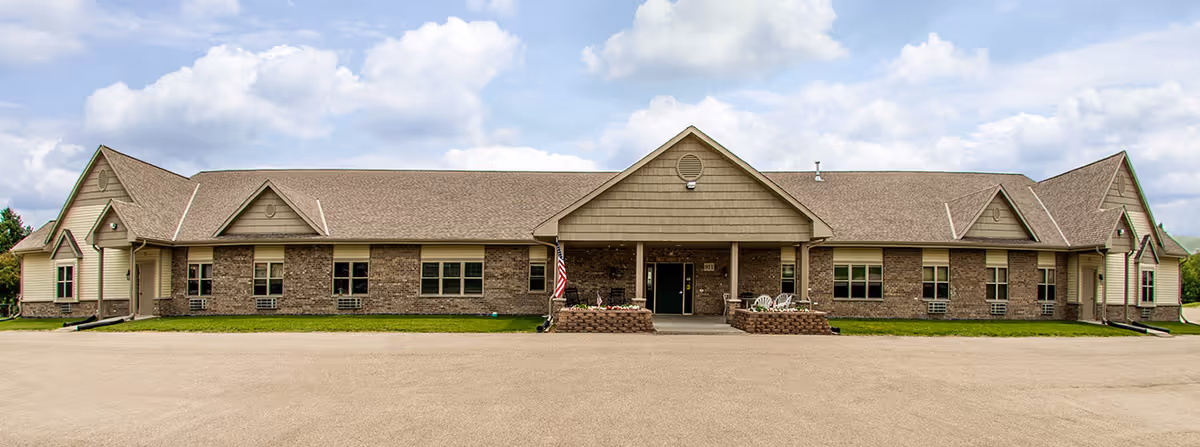 Front exterior view of a single-story senior living facility building with a brick facade, multiple windows, a central entrance with a covered porch, and an American flag displayed near the entrance under a partly cloudy sky.