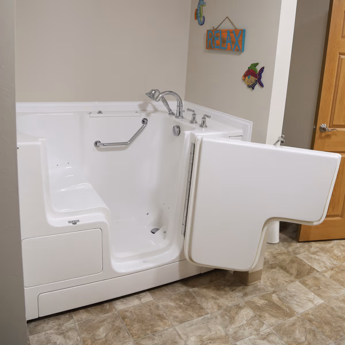 A white walk-in bathtub with an open door, chrome faucet, and grab bar inside. The tub is installed in a bathroom with beige walls, a wooden door, and decorative wall hangings including a sign that says 'RELAX' and colorful fish. The floor has beige and brown tiles.