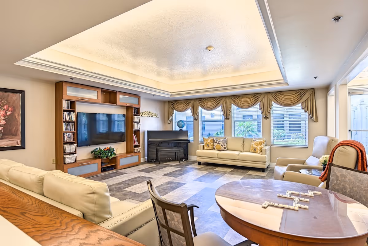 A bright and spacious living room with a large flat-screen TV mounted on a wooden entertainment center, a piano in the corner, two beige sofas with decorative pillows, and a round table with chairs and dominoes on it. Large windows with beige valances let in natural light.