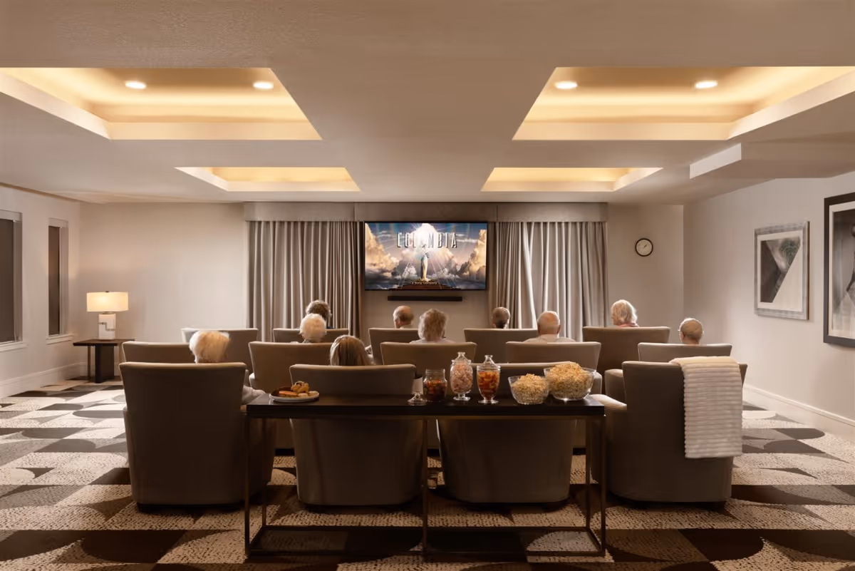 A group of elderly people seated in a cozy, well-lit room watching a movie on a large flat-screen TV mounted on the wall. The room has comfortable armchairs arranged in rows, a table with snacks including popcorn and drinks, patterned carpet, and soft lighting from recessed ceiling fixtures.
