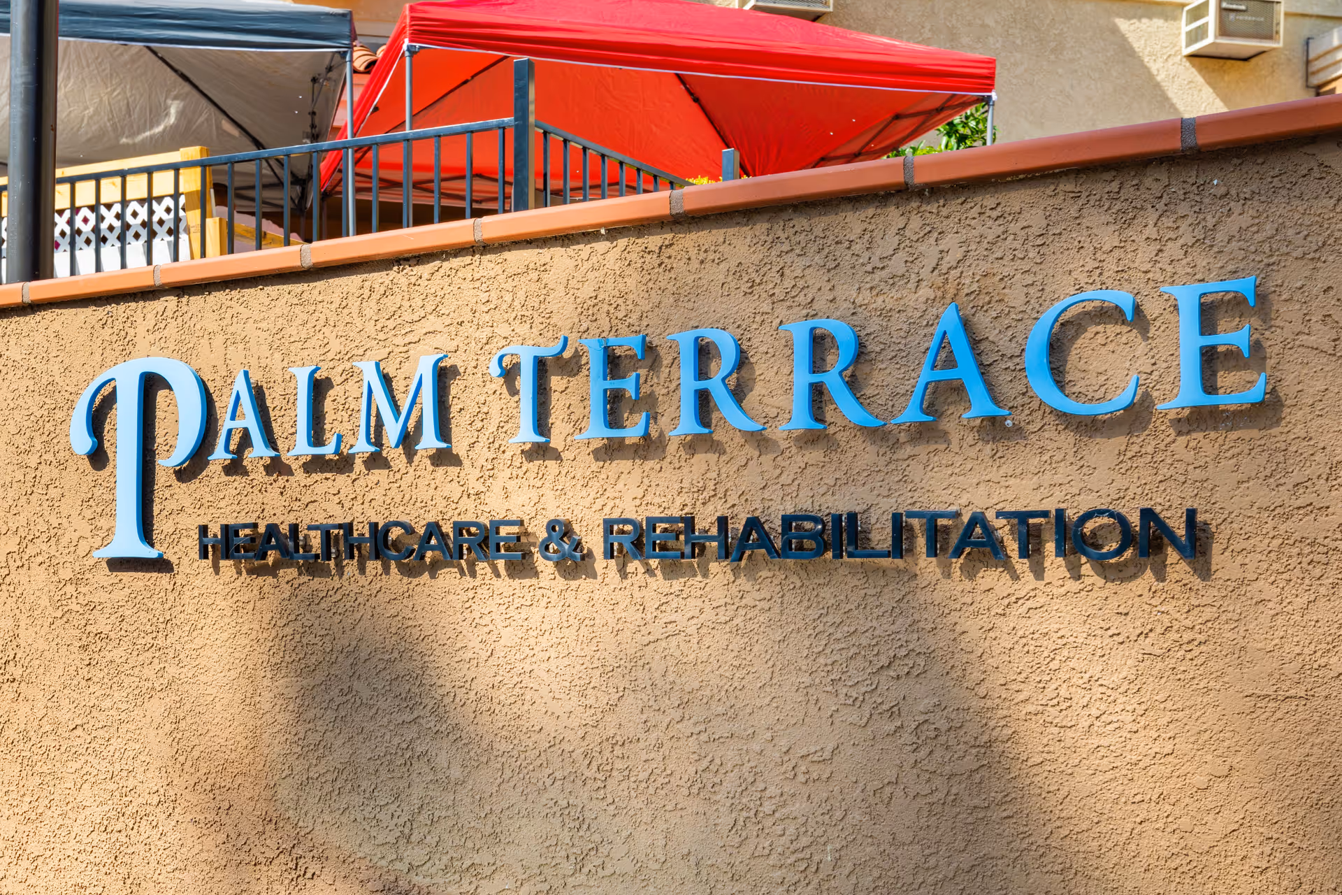 Sign on a stucco exterior wall reading "Palm Terrace Healthcare & Rehabilitation" with red umbrellas and a railing visible above.