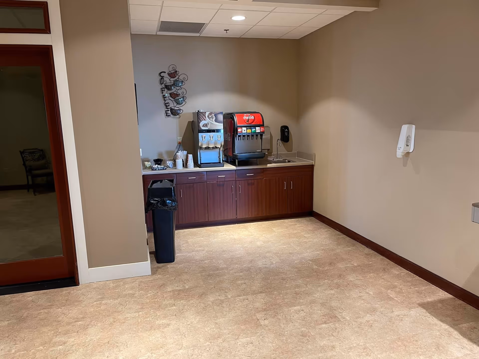 Beverage station in a common area with a soda fountain, coffee dispenser, sink, countertop and trash can.