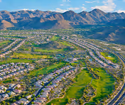 Aerial view of a residential community with houses arranged in curved rows surrounded by green spaces and a golf course, set against a backdrop of rugged mountains under a partly cloudy sky.