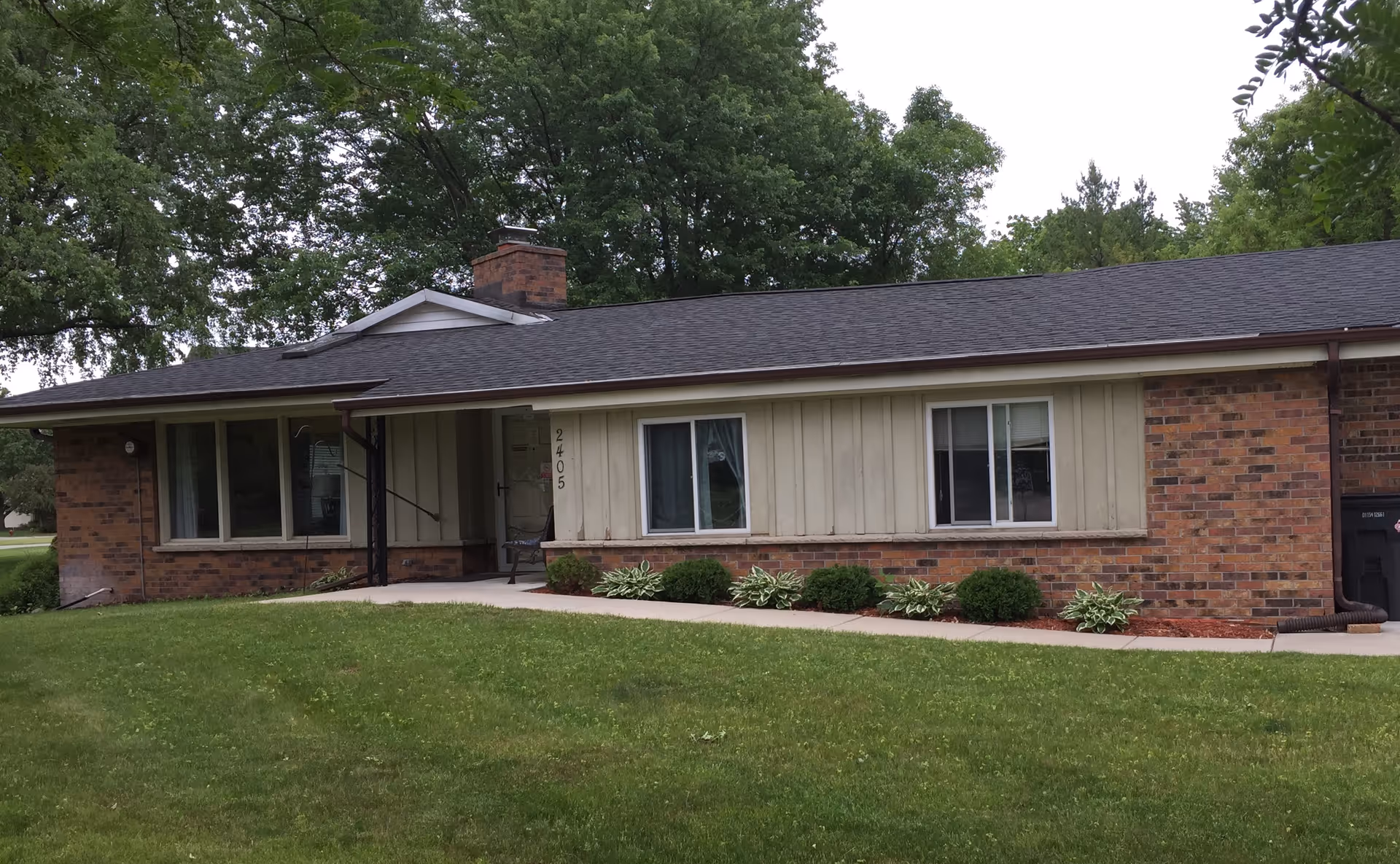 Single-story brick and siding residential building with a front porch, two windows, a chimney, and a well-maintained lawn with shrubs and trees in the background.