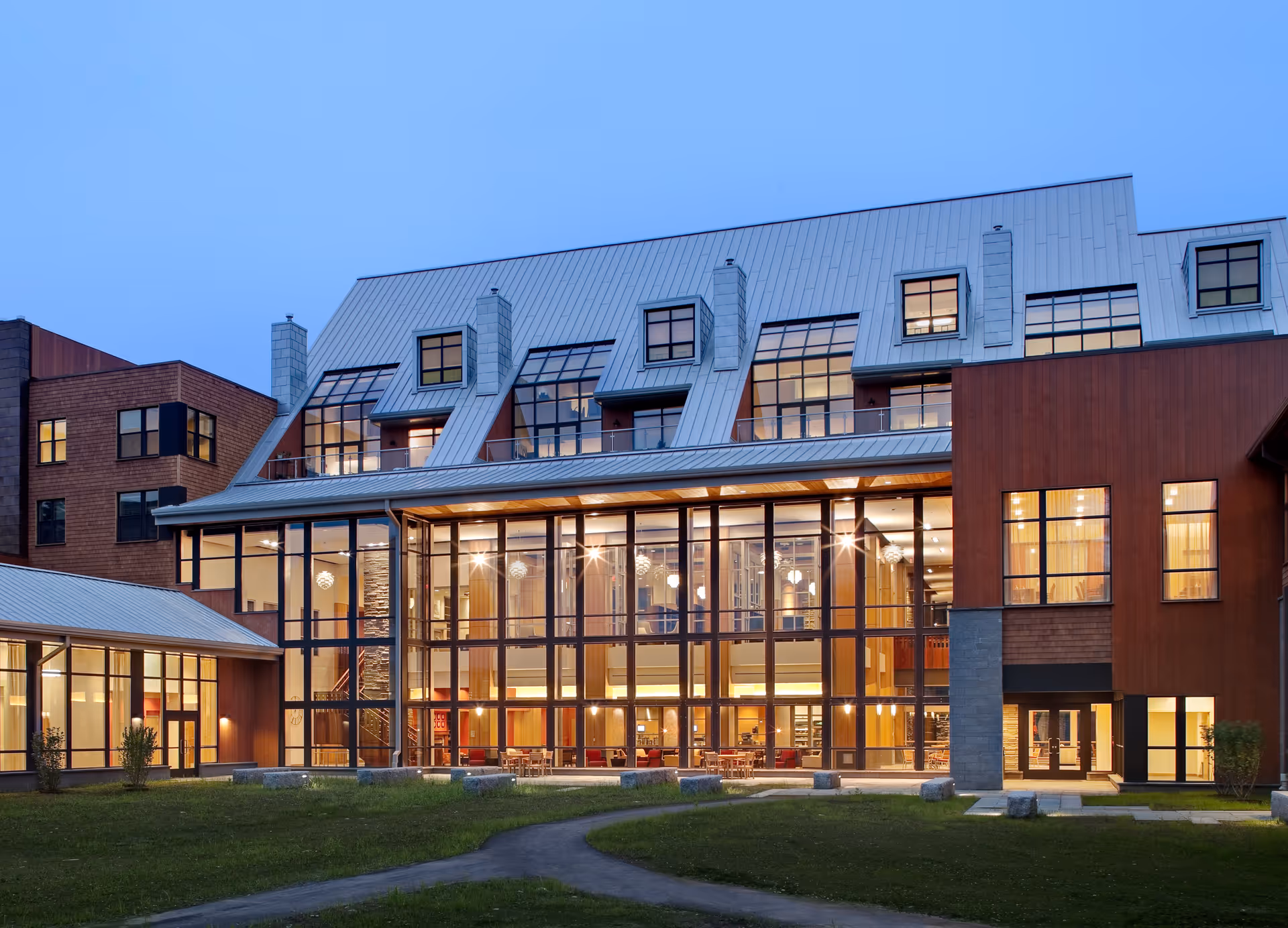 Exterior view of a modern senior living facility building at dusk with large glass windows showing warm interior lighting and a well-maintained lawn in the foreground.