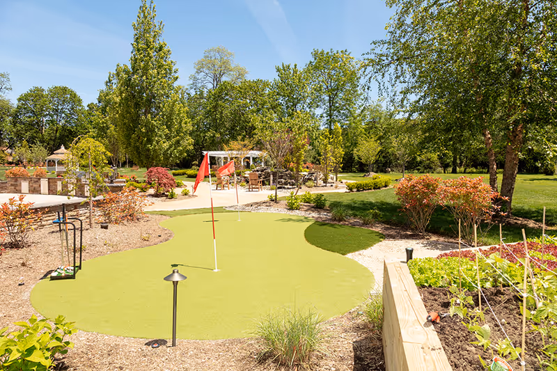 A vibrant garden area featuring a putting green with flags, surrounded by lush greenery and colorful plants, creating a serene outdoor space.