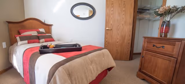 A cozy bedroom with a single bed featuring a striped red, brown, and beige comforter and matching pillows. A black tray with a small decorative item is placed on the bed. There is a wooden dresser with a vase of flowers on top, a wooden door leading to a closet with hanging clothes, and an oval mirror on the wall above the bed.